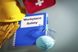 Workplace safety handbook manual and equipment. A red first aid kit in the back with a white cross on it, a hard hat on top of a pair of work gloves, a manual with the words "Workplace Safety" written in black on the cover. There is a light blue mask and a clear pair of safety glasses on top of the manual.